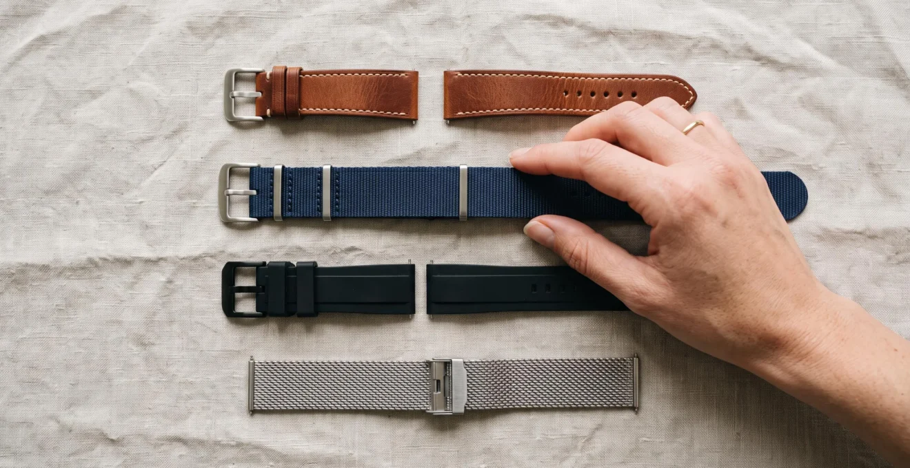 Quatre bracelets de montre de matières différentes posés côte à côte, une main s'apprêtant à choisir l'un d'eux.